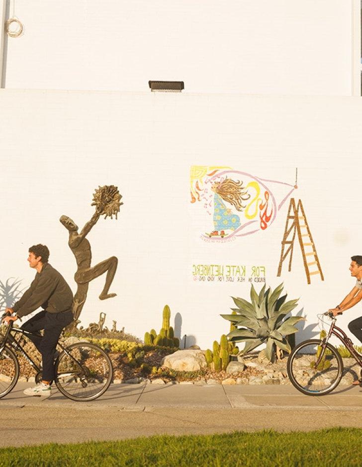 three students ride bikes past the front of mead hall